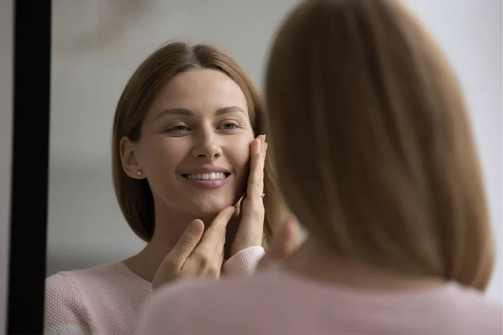 Woman looking in the mirror and gently touching her cheek after cosmetic treatment.