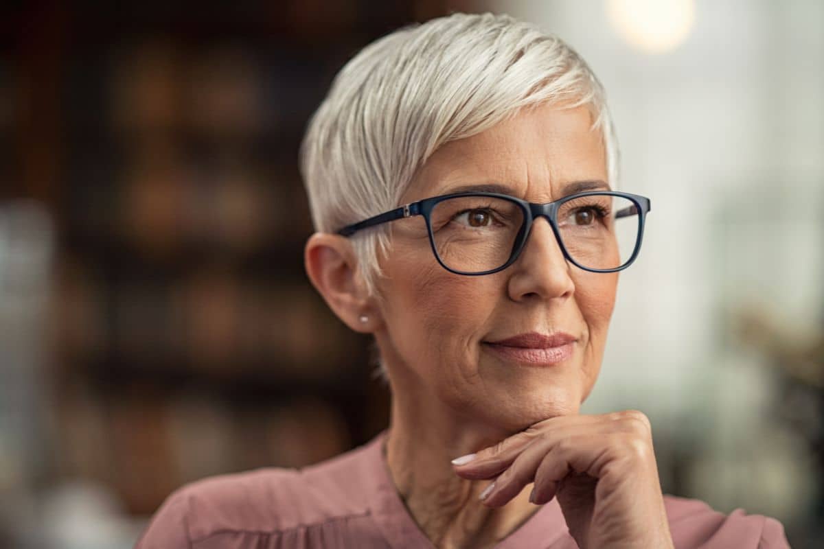 Elderly woman with short white hair and glasses reflecting calmly, representing facial skin cancer reconstruction concerns.