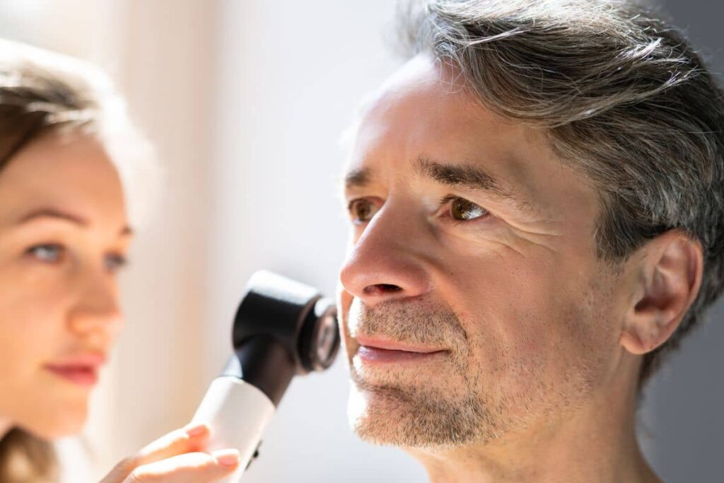 Doctor using dermatoscope to examine man’s face closely for signs of recurring or new skin cancer.