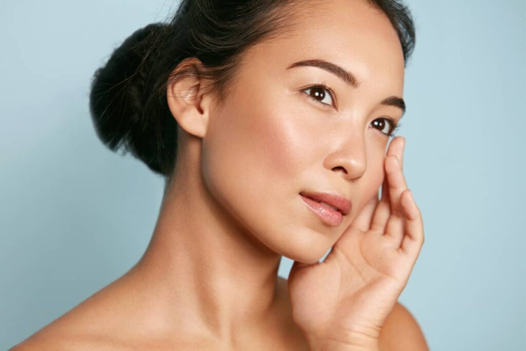 A close-up of an Asian woman with her hair in a bun, looking upward and touching her cheek to showcase her elegant facial profile and contoured nose.