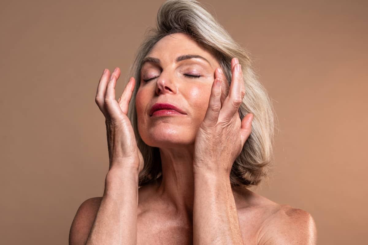 A woman with silver hair and eyes closed gently touches her face with both hands, showcasing a smooth and healthy complexion.