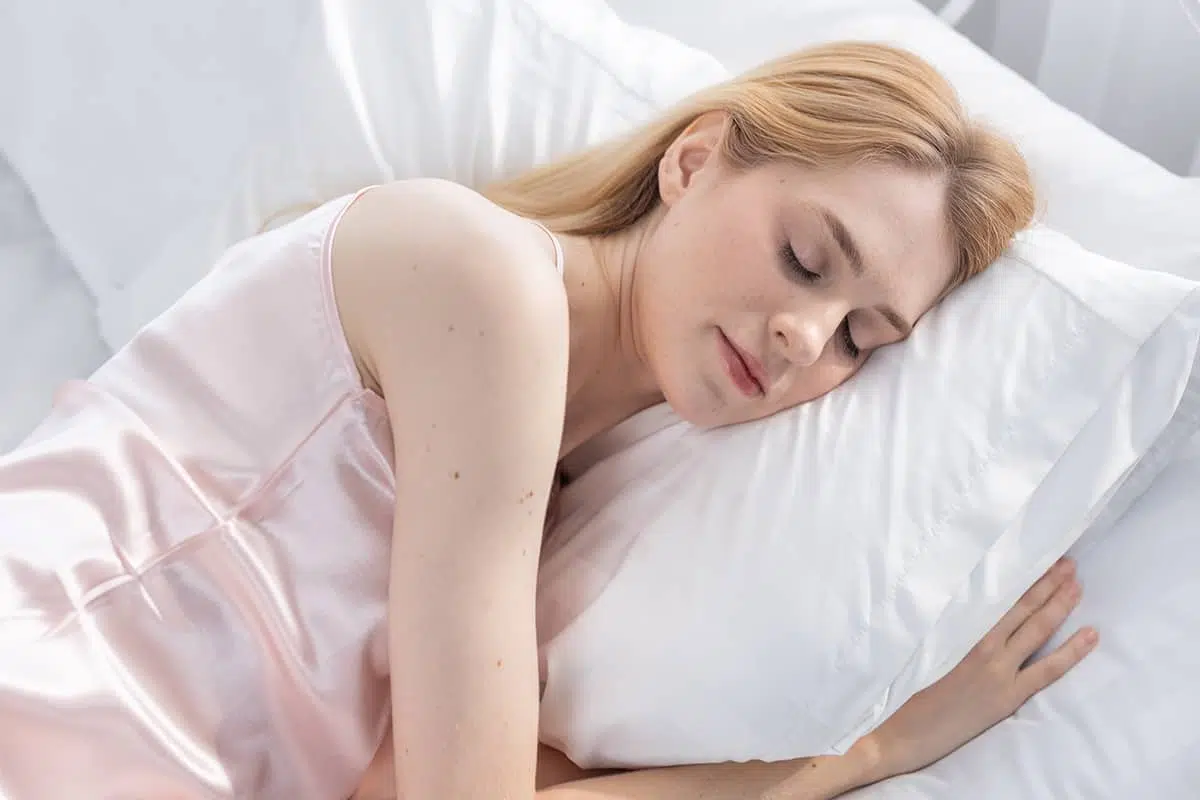 A blonde woman in a pink satin top sleeping peacefully on her side on a plush white pillow.