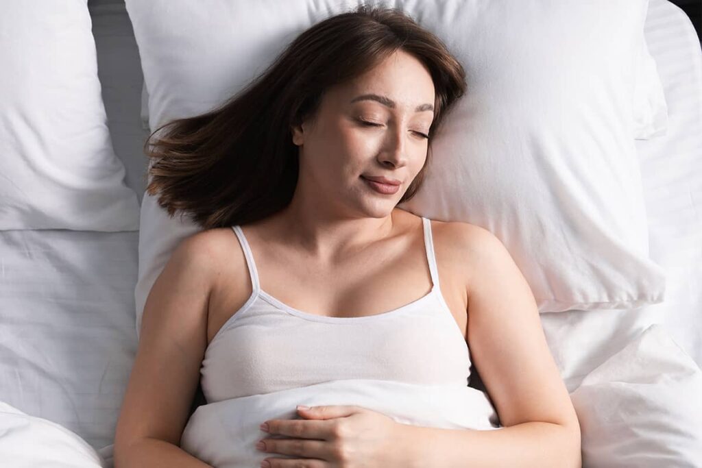 An overhead view of a brunette woman sleeping comfortably on her back on a crisp white pillow.